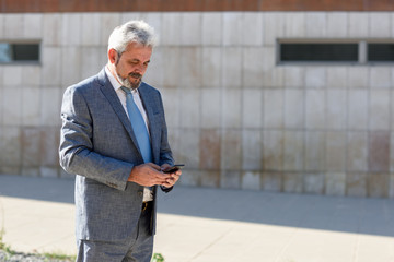 Senior businessman texting with smartphone outside of modern office building.