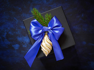Christmas gift Stylish black on blue background. Decorated with a bow of blue ribbon, gold Christmas tree toy and branches of fir.