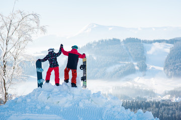 Couple snowboarders high fiving each other, standing on top of a snowy mountain, observing stunning...