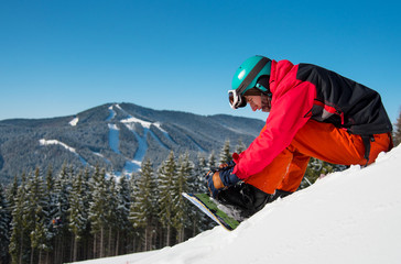 Shot of a snowboarder sitting on the snowy slope, preparing for riding downhill on a beautiful sunny winter day. Blue sky, mountains, forests, ski slopes on the background. copyspace sports concept