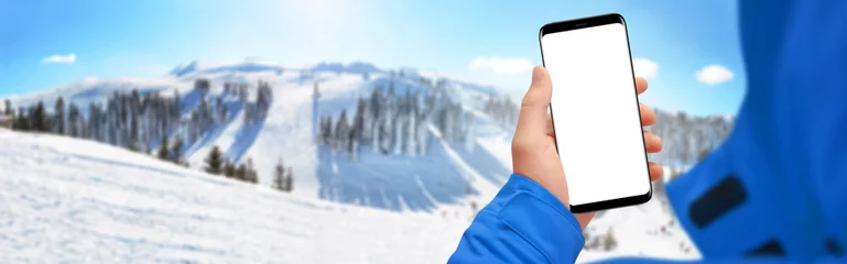 Poster Man hand holding modern smartphone with isolated screen, snowy mountain in background © nikolas_stock