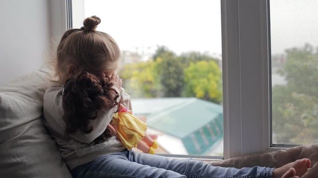 Beautiful young girl with doll sitting on a window sill