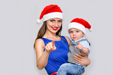 Fototapeta premium Young mom holding a baby and showing towards the camera.Both in Santa hats. New year and the fun will be soon. 