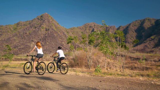 Two women on bicycles riding on rural dirt road in front of dry mountains, chicken runs away. Shot with Sony a7s and Atomos Ninja Flame on sunny summer day in Bali, Indonesia