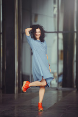 cheerful curly woman in blue dress and red shoes in the city business center, holiday shopping
