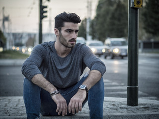 Attractive young man portrait at night with city lights behind him in Turin, Italy, sitting on sidewalk curb