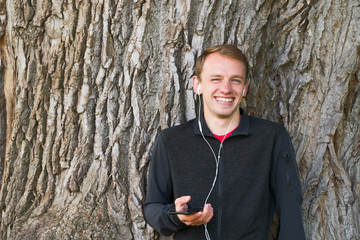 Young smiling man with sporty clothes listening to music with headphones from her smart phone and relaxing at the park, sunny day