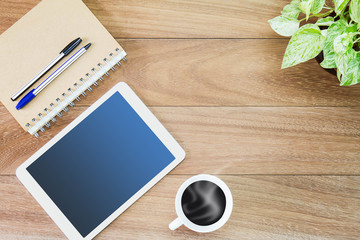 Tablet computer with hot coffee cup, green plant pot, notebook and pens on wooden desk