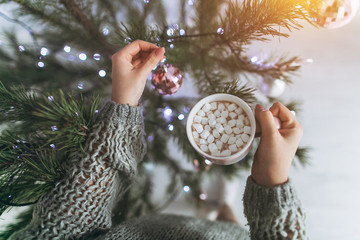 A girl drinks cacao with marshmallo and decorates a Christmas tree.