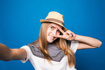 Fototapeta premium Trendy beautiful hipster girl taking selfie with victory sign on mobile phone against a blue wall.