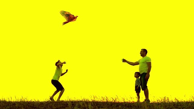 Happy family father and children running in the meadow with a kite in summer at sunset in a slow motion. Family outdoors, the sky is yellow. Yellow background