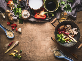 Vegetarian stir fry cooking preparation on wooden background with various vegetables, wok, coconut milk, seeds and kitchen utensils, top view, frame. Asian cuisine. Healthy eating and food concept