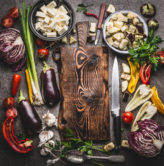 Various vegetables cooking ingredients around wooden cutting board with knife on rustic background with bowls. Vegetarian cooking preparation for tasty dishes, top view. Healthy eating concept