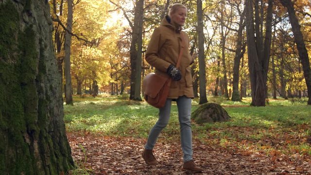 Thoughtful woman walking in the park appearing from old oak tree. Slider dolly shot. Sunny afternoon in natural park in autumn. Colorful leaves inder the trees.