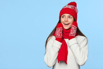 Surprised happy woman looking sideways in excitement. Christmas girl wearing knitted warm hat and scarf, isolated on blue background
