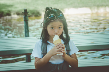 Asian little cute girl sitiing on wooden bench and eating ice cream at the park in vintage style.