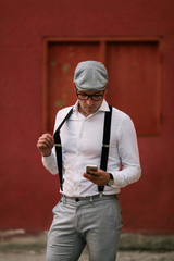 Naklejka premium Fashionable retro dressed man with cap, suspenders and eyeglasses standing on city street and using smart phone.