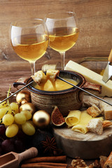 Gourmet Swiss fondue dinner on a winter evening with assorted cheeses on a board alongside a heated pot of cheese fondue with two forks dipping bread and white wine behind in a tavern or restaurant