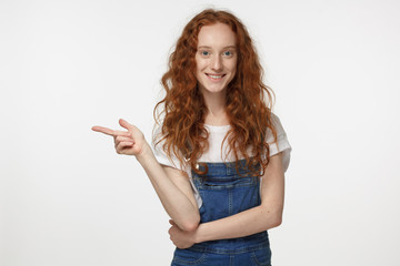 Beautiful young redhead curly female in jean jumpsuit pointing left with finger isolated on gray background