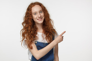 Casual dressed young hipster redhead curly female pointing right with his finger isolated on gray background
