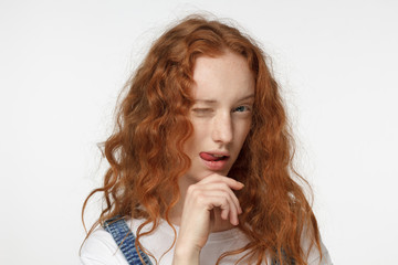 Pretty redhead curly girl smiling and winking, looking at camera isolated on gray background