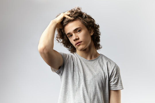 Fashionable handsome young European male model dressed in grey t-shirt posing in studio for commercial photo shooting, looking at camera and adjusting his voluminous hair, advertising shampoo