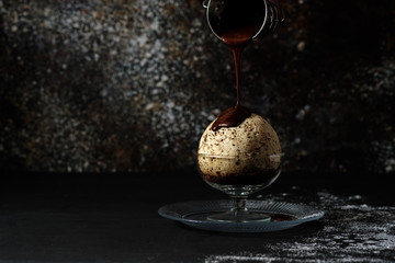 A large bowl of ice cream in a glass cup is poured with dark chocolate.