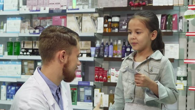 The chemist is sitting next to a little girl. She is unwell and he gives her the necessary pills. She smiles and shakes his hand. Mom and daughter are in the pharmacy