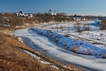 Early spring in Suzdal/City landscape of Suzdal. River, ice-bound, In the flood plain lies snow. On the slope of the hill, the snow melted and last year's grass is visible.Suzdal.Golden Ring of Russia