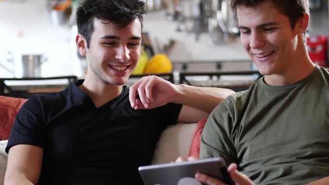 Two casual young men friends relaxing on sofa at home sharing information and using tablet computer