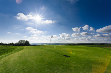 View of a green golf course, hole and flag on a bright sunny day. Sport, relax, recreation and leisure concept. Summer landscape with sunbeams