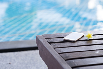 Wooden chair with smart phone and flower near swimming pool