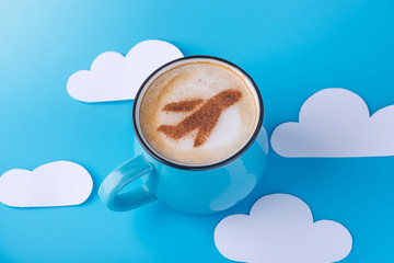 Cup of cappuccino with a picture of the plane on the foam. Blue sky background with paper clouds. Concept of travel