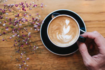 Top view: a man's hand holds a cup of coffee with flowers nearby on a wooden surface.