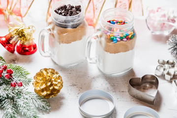 Christmas homemade gift concept, ingredients for preparation chocolate and rainbow cookies in jar, marshmallow in dish, metallic cutting forms for cookies and decorations on kitchen table