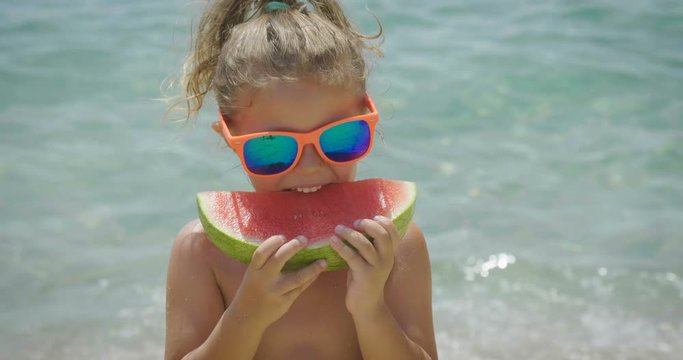 Portrait of little girl eats fresh watermelon at sea, smiling happily, wearing sunglasses, in swimsuit, background of sea blue water. Concept: fresh fruits, vacations, summer, children, daughter, kids