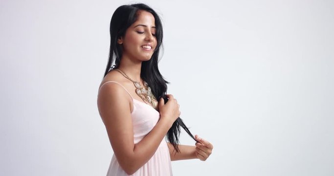 Smiling happy Indian girl in light pink dress and heavy necklace playing with her gorgeous black straight hair on white background