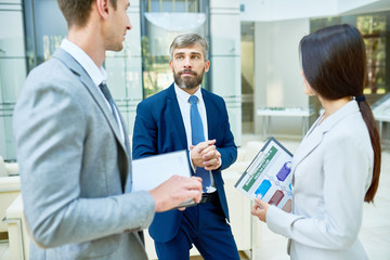 Portrait of three business people meeting in hall of modern office building talking and discussing work during break, focus on successful bearded businessman in center