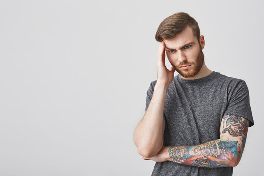 Good-looking tattooed mature sad man with beard and fashionable hairstyle massaging forehead with hand, looking aside painful expression, being exhausted with strong headache.
