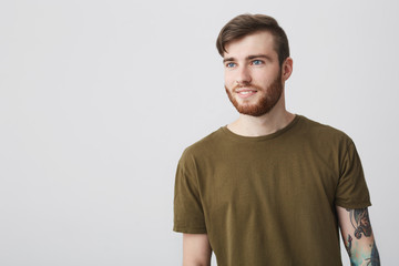 Fototapeta premium Positive emotions. Close up portrait of good-looking mature european bearded man with blue eyes and tattoo sleeve in olive casual t shirt looking aside with inspired and satisfied expression,