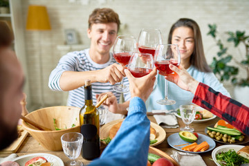 Group of friends enjoying dinner together sitting at big table with delicious food , focus on clinking wine glasses in celebration of holiday
