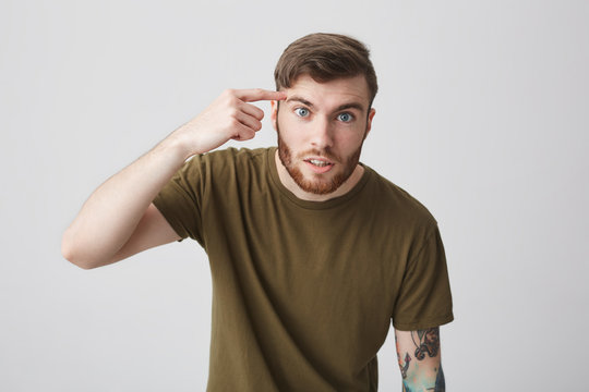 Are you stupid. People's emotions. Close up of handsome negative bearded male student with short hair and colorful tattoo on arm in brown casual t-shirt looking in camera with angry expression