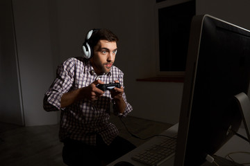 Portrait of young funny bearded man in headphones wearing checked shirt playing computer games on personal computer with controller in night. Gaming, cyber sport.