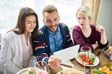 Portrait of three young people smiling happily video chatting from difital tablet at dinner table during holiday celebration