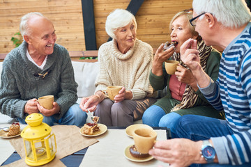 Group of elderly men and women warming themselves with hot tea and enjoying delicious cake while having gathering at cozy small patio - obrazy, fototapety, plakaty