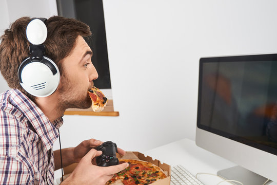 Close up of young good-looking unshaven man with short hair in casual shirt playing computer games with controller in headphones, eating pizza, spending free time talking and playing with friends