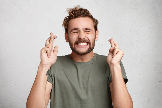 Cheerful bearded man keeps fingers crossed, smiles broadly and closes eyes, makes desire wish. Positive male student believes in victory or success on exam. Feelings, attitudes and reactions