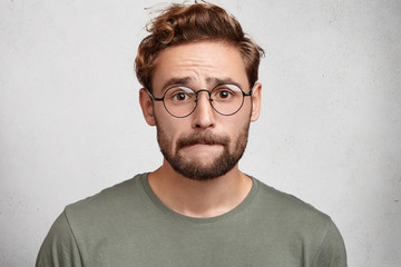Worried bearded guy in eyewear, bites lower lip, anticipate important decision or feels nervous...
