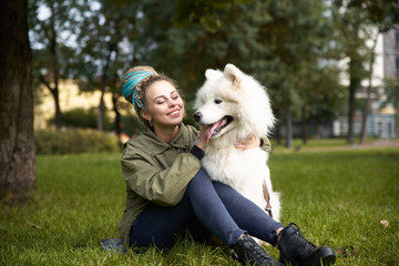 cheerful young girl in a green coat and dreadlocks and piercings sitting on the grass with his snow-white Samoyed dog in the autumn Park