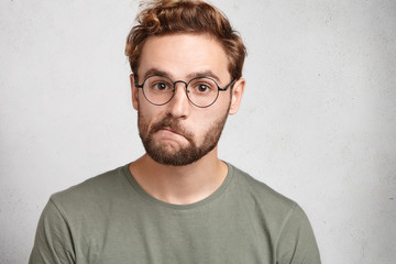 Indoor shot of doubtful bearded man in spectacles, curves lips, looks directly into camera, hesitates what answer to give. Uncertaing bearded male student at final exam, frowns face in dubiety